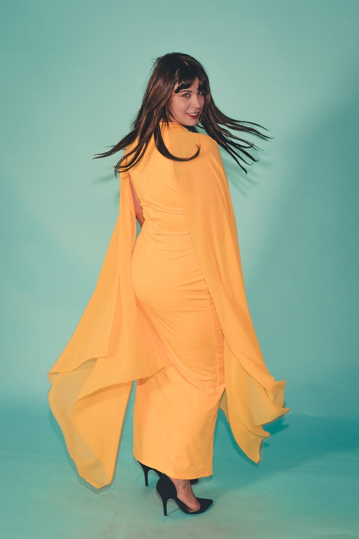Stylish adult woman in a vibrant yellow dress poses gracefully indoors against a pastel background.