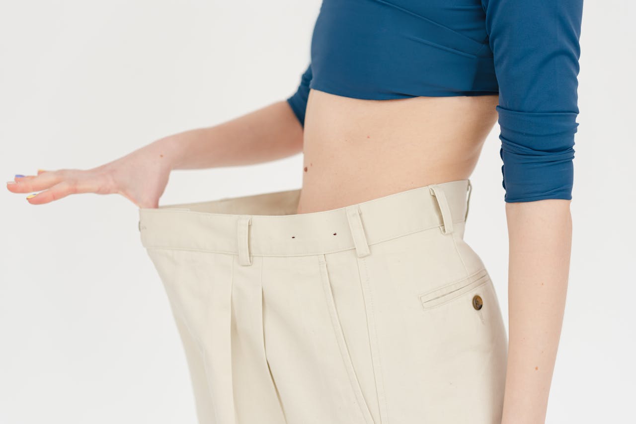 A woman demonstrates significant weight loss by wearing oversized pants indoors.
